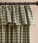 A close-up of a pleated curtain panel with a green and beige gingham plaid pattern, hanging from a dark brown metal rod against a light beige wall.