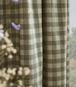 A close-up of a window curtain featuring a classic gingham check pattern in muted olive green and cream, with soft-focus baby's breath flowers in the foreground.