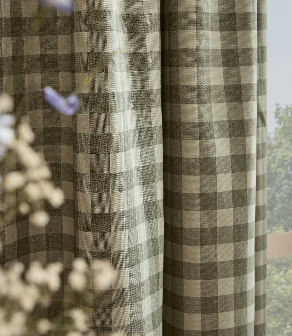 A close-up of a window curtain featuring a classic gingham check pattern in muted olive green and cream, with soft-focus baby's breath flowers in the foreground.