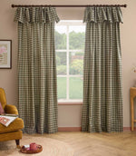 A pair of green and beige gingham check curtain panels with matching ruffled valances hanging on a dark wood rod in a styled room with a window, wooden floor, and a mustard yellow armchair.