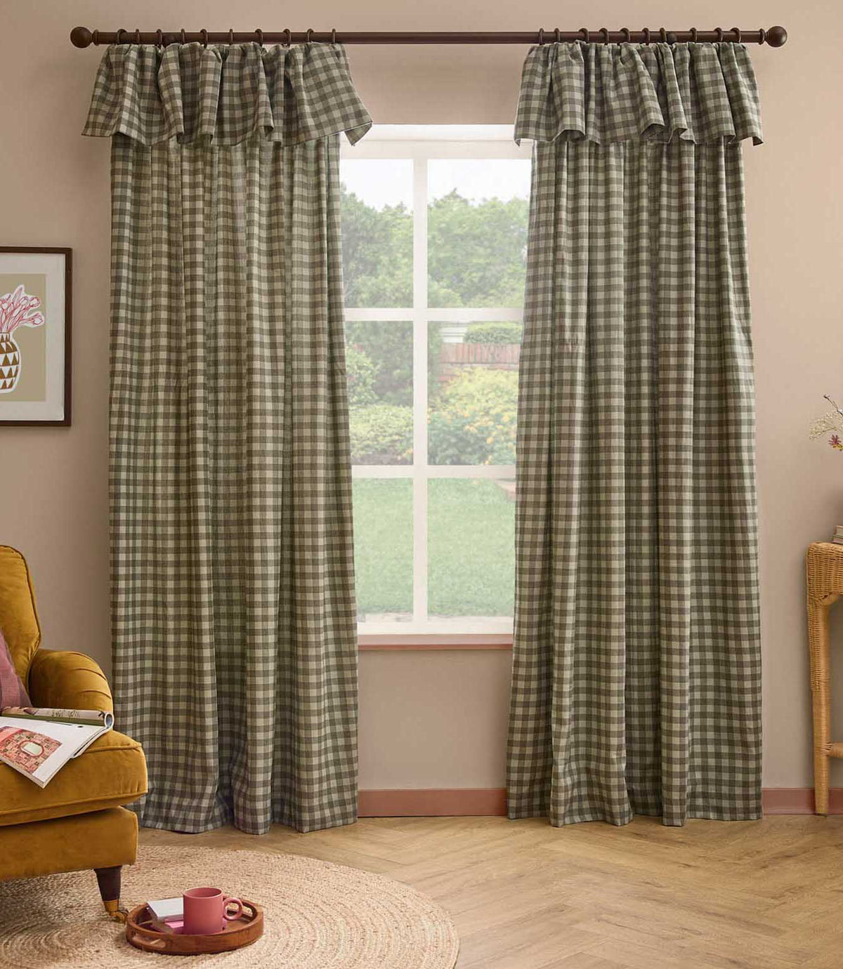 A pair of green and beige gingham check curtain panels with matching ruffled valances hanging on a dark wood rod in a styled room with a window, wooden floor, and a mustard yellow armchair.