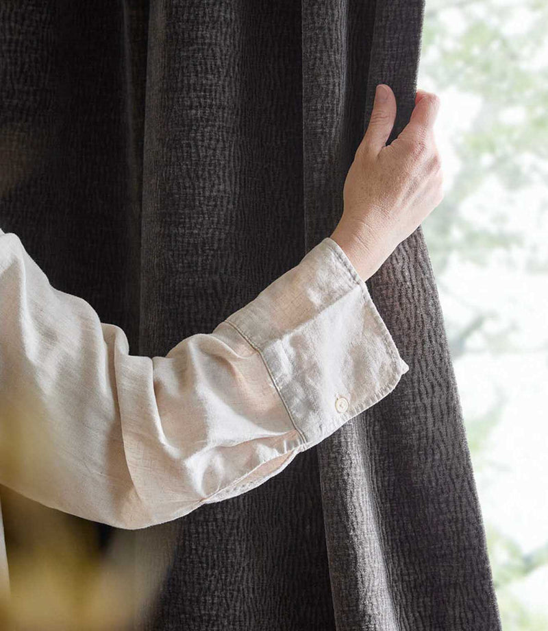 A person gently holds a charcoal grey Heath washed velvet curtain beside a bright window, showing its soft pile and subtle woven texture.