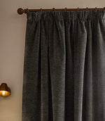 Close view of charcoal grey Heath washed velvet pencil pleat curtains hanging from a dark wooden pole beside a warm wall light.