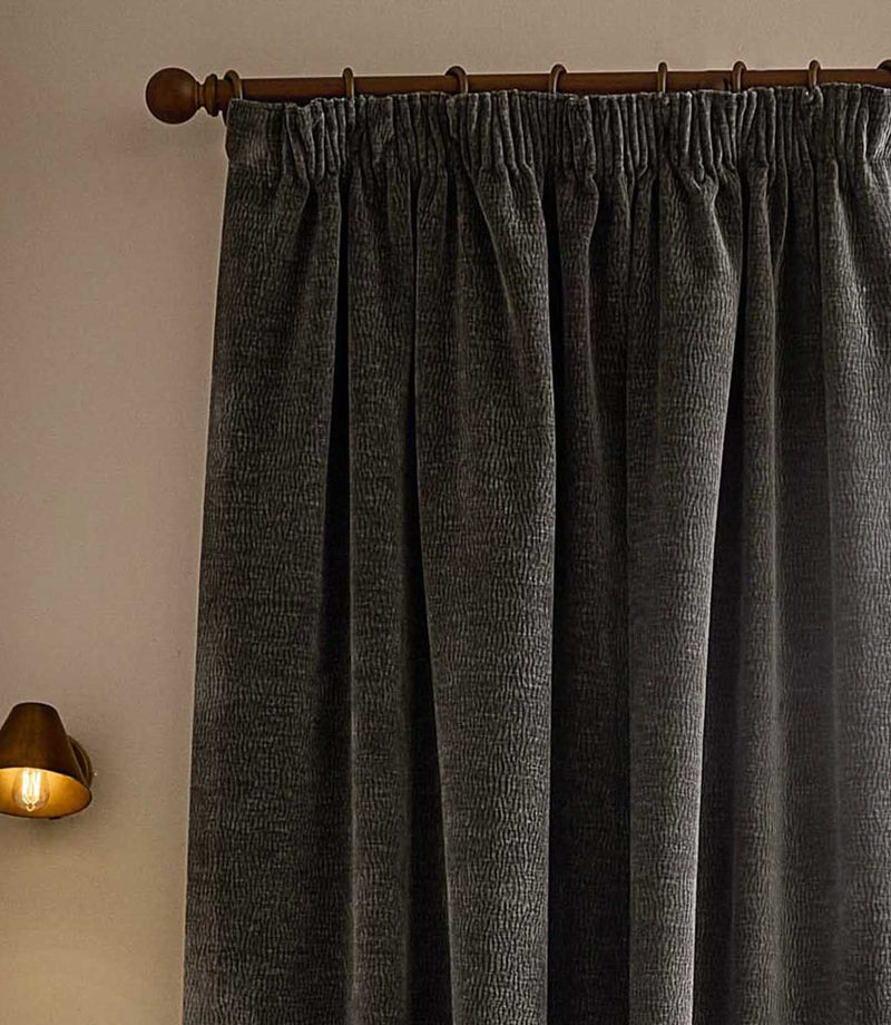 Close view of charcoal grey Heath washed velvet pencil pleat curtains hanging from a dark wooden pole beside a warm wall light.