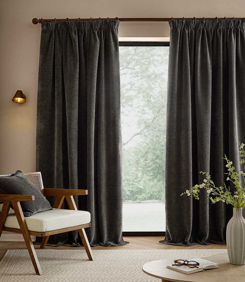 Floor-length charcoal grey Heath washed velvet curtains frame a large window in a neutral living room with a wooden armchair and matching cushion.