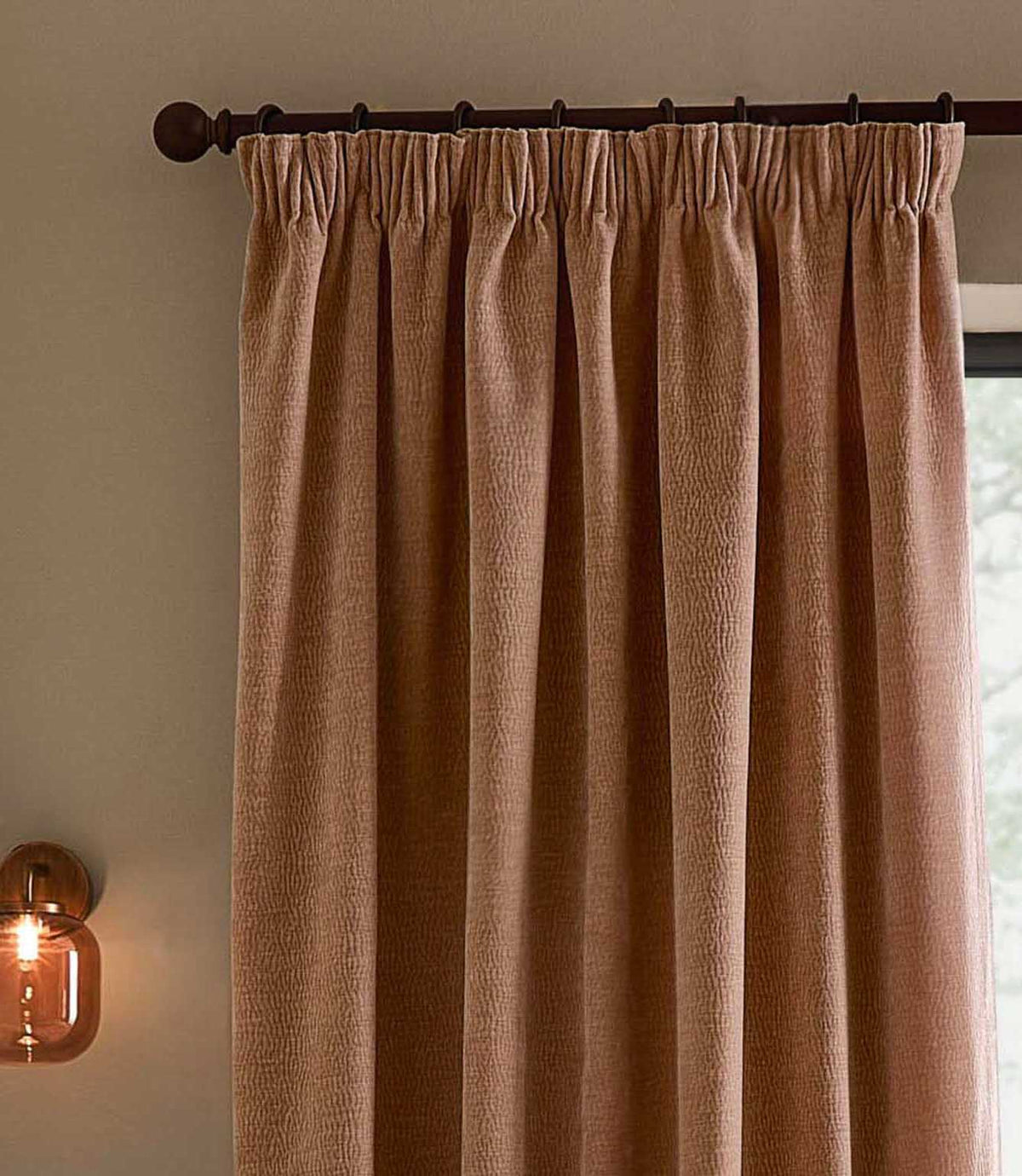 Close view of a soft terracotta-pink Heath Washed Velvet pencil pleat curtain in Cotta, hanging from a dark wooden pole beside an amber wall light.
