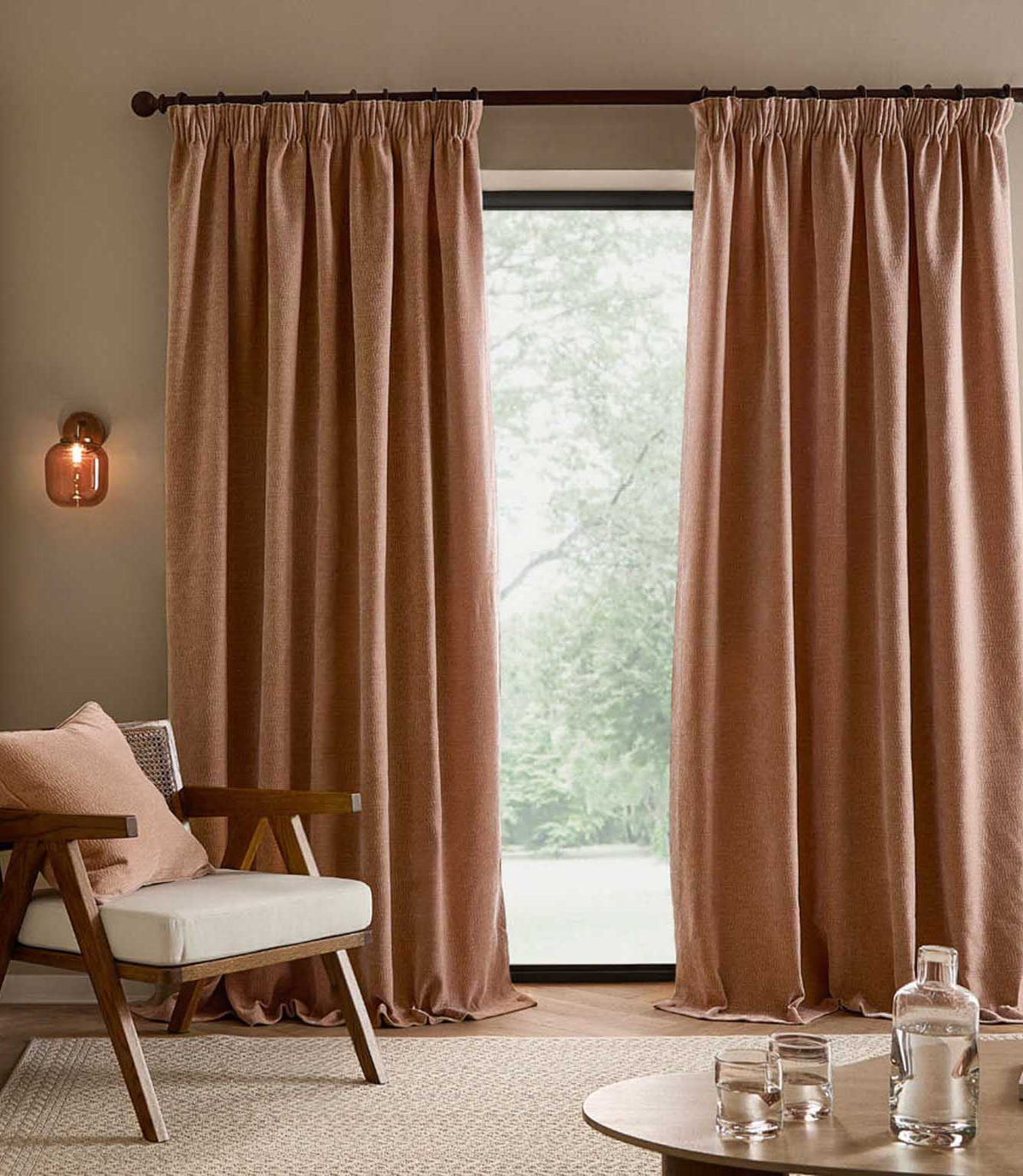 Floor-length Heath Washed Velvet curtains in soft terracotta-pink Cotta frame a large window in a neutral living room with a wooden armchair and matching cushion.
