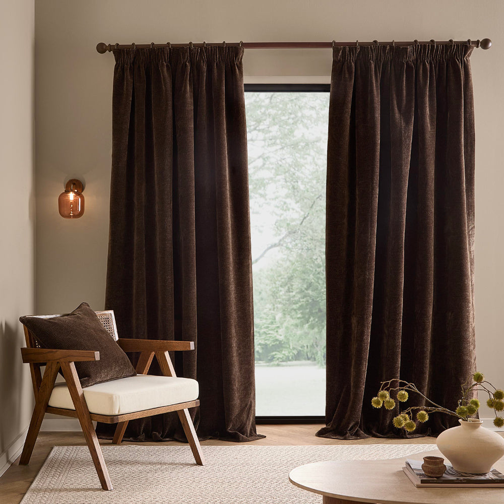 Full-length espresso brown washed velvet curtains framing a large window in a neutral living room, styled with a wooden armchair and coordinating cushion.
