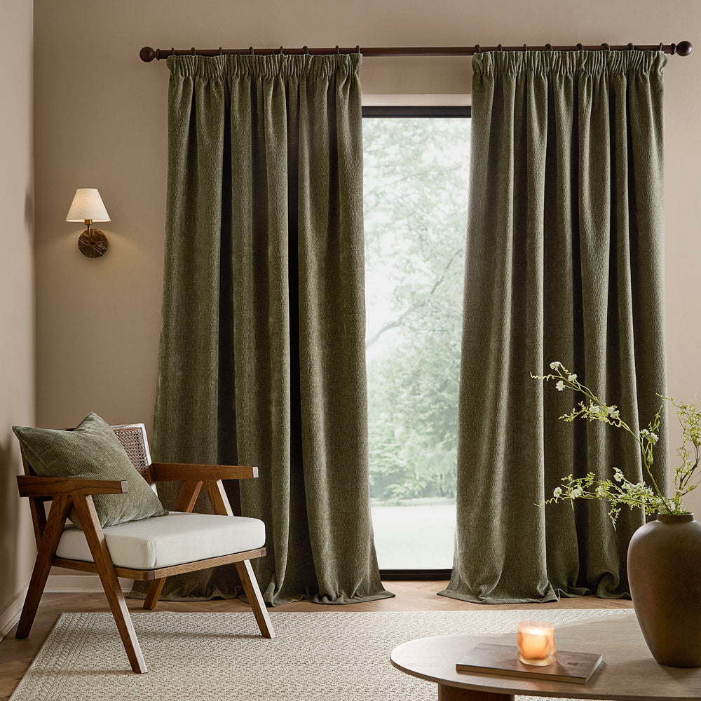 Full-length eucalyptus green washed velvet curtains framing a large window in a neutral living room, styled with a wooden armchair, coordinating cushion and natural accessories.