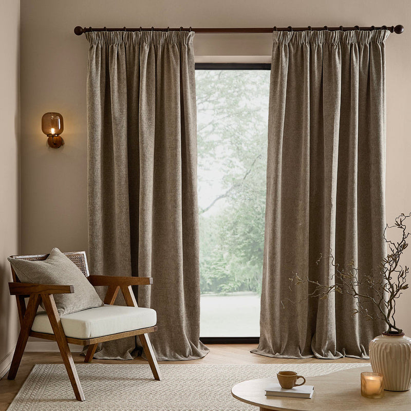 Full-length greige washed velvet curtains framing a large window in a neutral living room, styled with a wooden armchair, coordinating cushion and natural accessories.