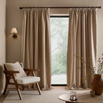 Full-length natural linen beige washed velvet curtains framing a large window in a neutral living room, styled with a wooden armchair, coordinating cushion and natural accessories.
