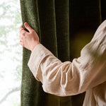 Close-up of a person holding an olive green washed velvet curtain beside a window, showing the soft drape, rich pile and distinctive textured finish.