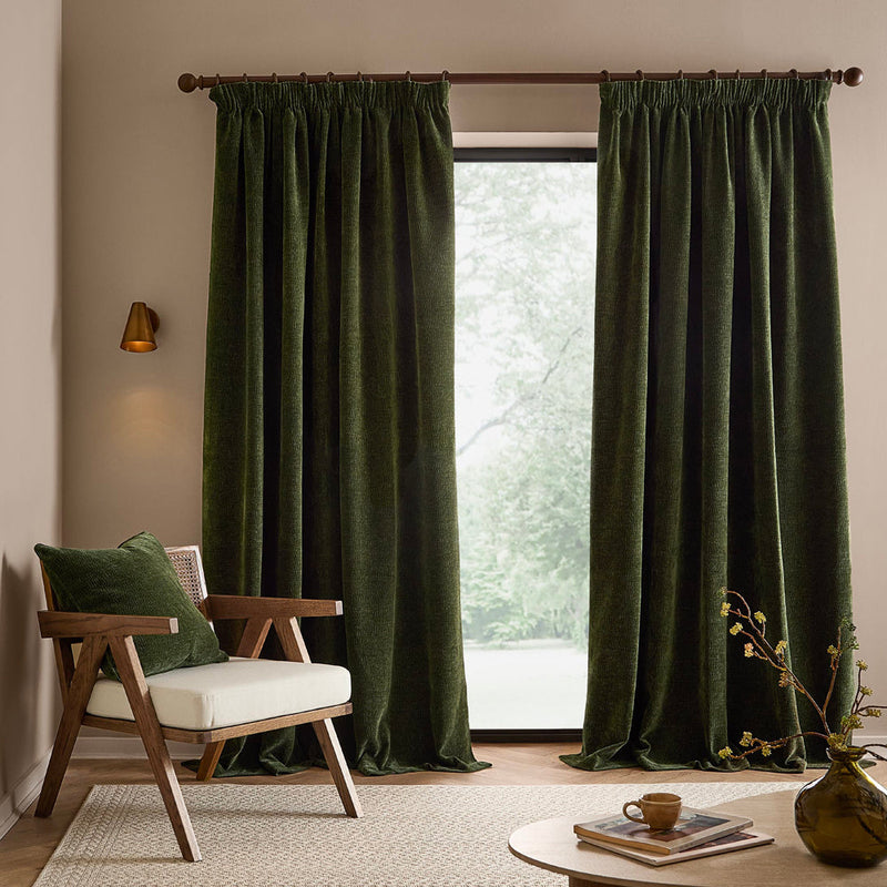 Full-length olive green washed velvet curtains framing a large window in a neutral living room, styled with a wooden armchair, matching green cushion and natural accessories.