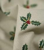 Detailed close-up of Holly and Berry Stone fabric showing a green holly sprig with red berries and delicate gold accents on a textured stone neutral background.