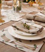 A styled festive table setting featuring beige placemats and napkins printed with green holly leaves and red berries, arranged with white plates, silver-toned cutlery, and a small sprig of greenery on a wooden dining table.