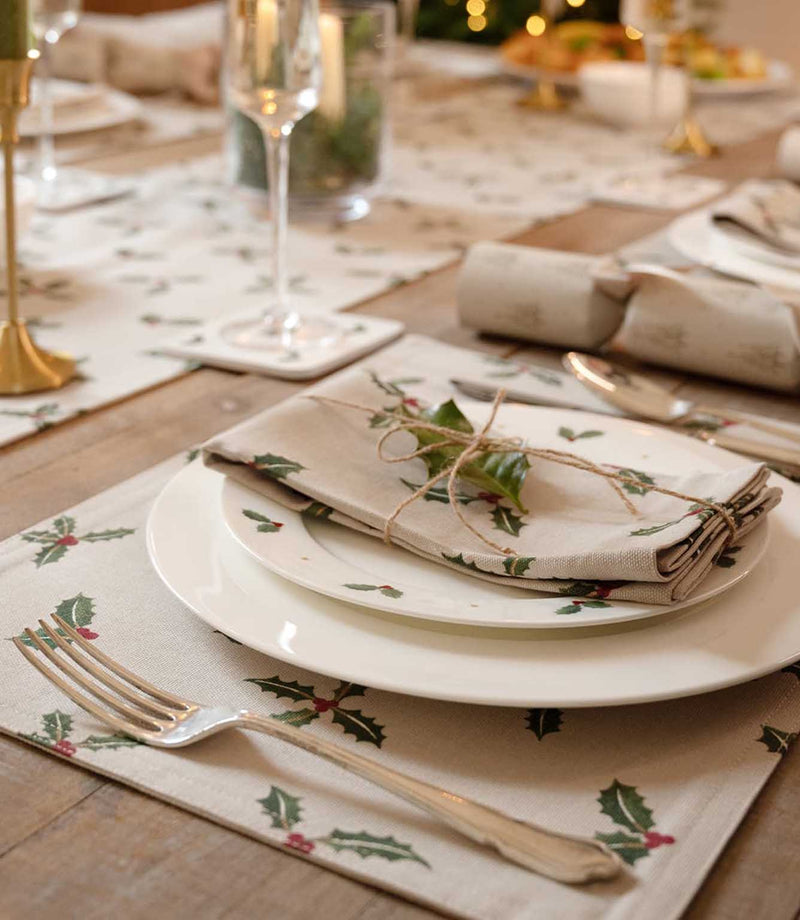 A styled festive table setting featuring beige placemats and napkins printed with green holly leaves and red berries, arranged with white plates, silver-toned cutlery, and a small sprig of greenery on a wooden dining table.