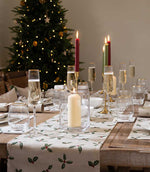 A festive Christmas table setting with a white table runner printed with green holly leaves and red berries, set on a rustic wooden dining table. The table is decorated with clear glass candle holders, lit taper candles in red and green, and gold-rimmed champagne glasses.