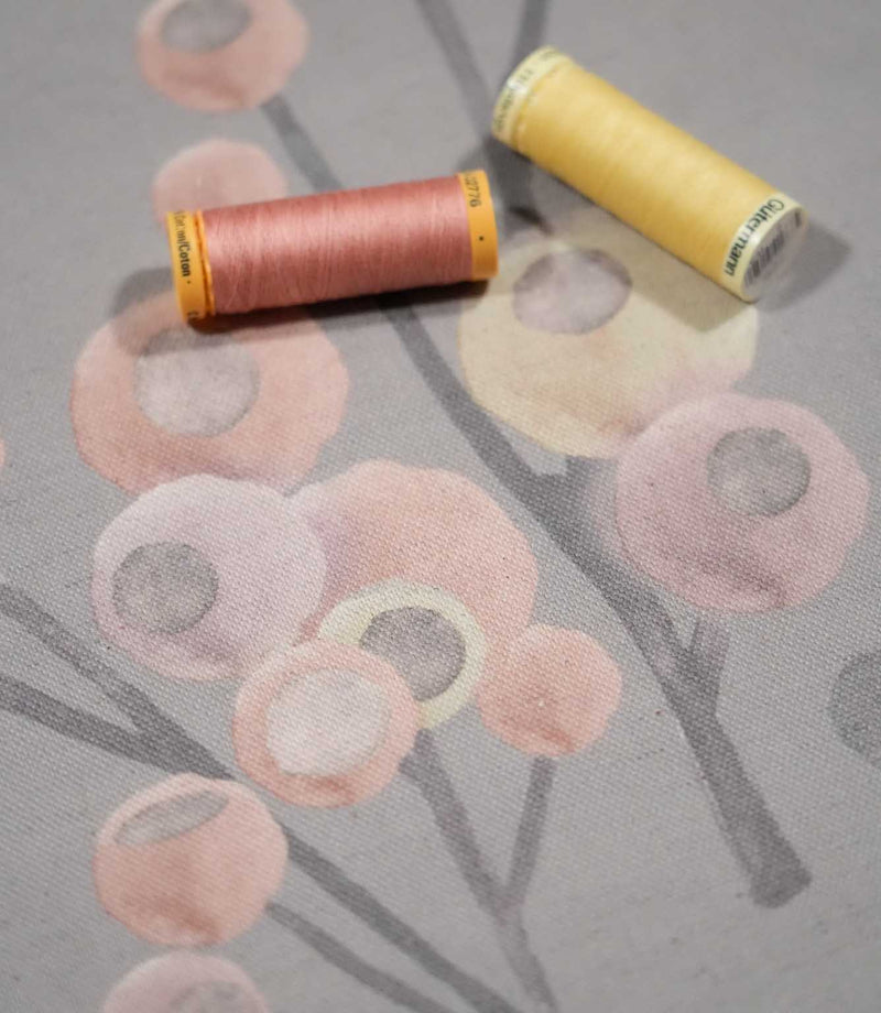 Ichiyo floral fabric laid flat with pink and yellow sewing thread spools, highlighting the soft grey background and delicate pastel botanical print.