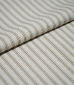 Close-up of folded beige ticking stripe fabric showing the woven texture, natural cream ground and fine neutral striped pattern.