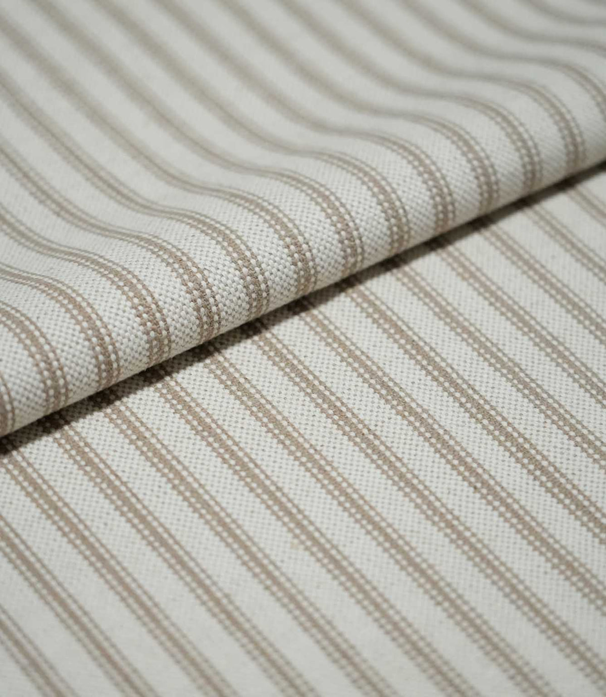 Close-up of folded beige ticking stripe fabric showing the woven texture, natural cream ground and fine neutral striped pattern.
