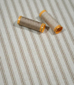 Beige ticking stripe fabric styled with neutral sewing thread, showing the cream woven ground and fine taupe-beige vertical stripes.