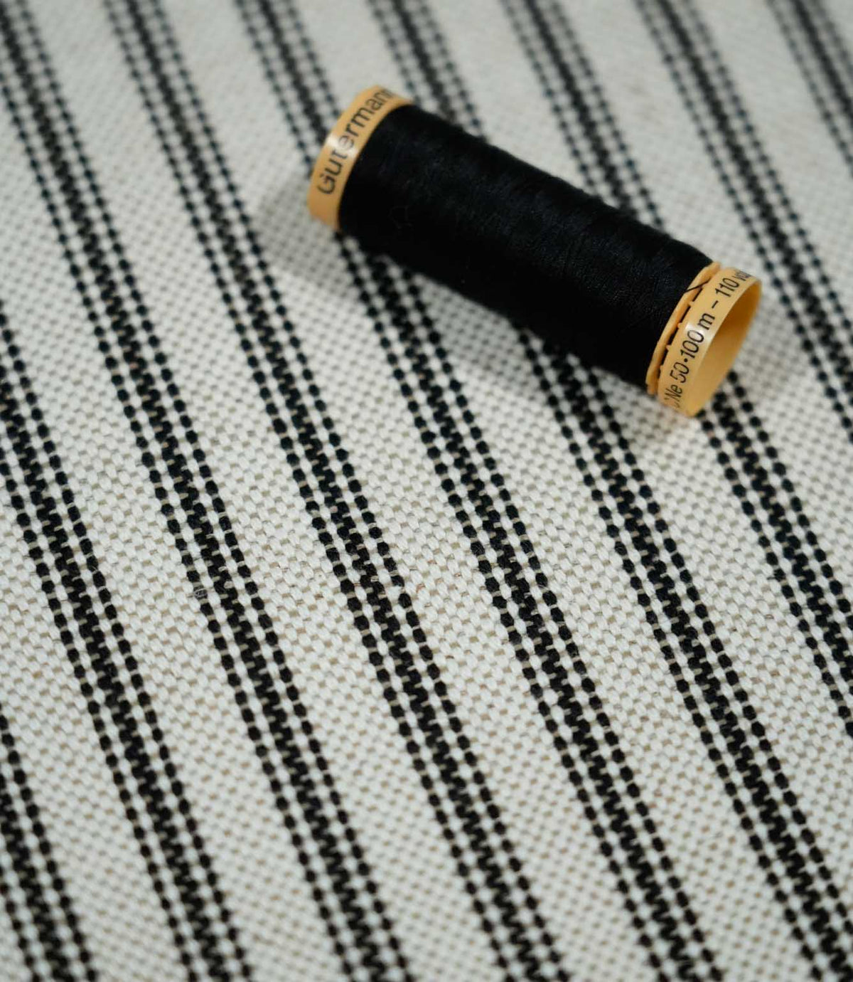 Close-up of black ticking stripe fabric with a black thread spool, highlighting the crisp black stripes and textured natural cream woven ground.