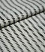 Folded black ticking stripe fabric showing the repeating narrow black stripes, natural cream background and tactile woven texture.