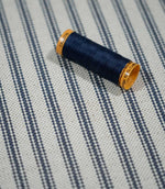Close-up of blue ticking stripe fabric with a navy thread spool, highlighting the woven navy stripes and natural cream textured background.