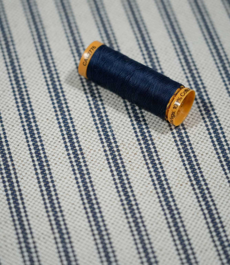Close-up of blue ticking stripe fabric with a navy thread spool, highlighting the woven navy stripes and natural cream textured background.
