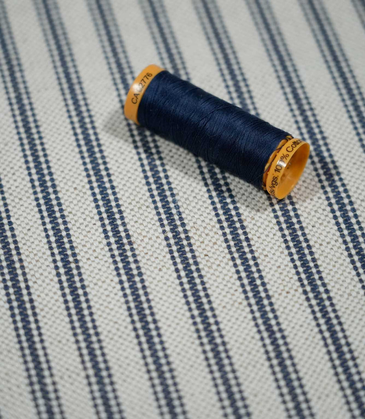 Close-up of blue ticking stripe fabric with a navy thread spool, highlighting the woven navy stripes and natural cream textured background.