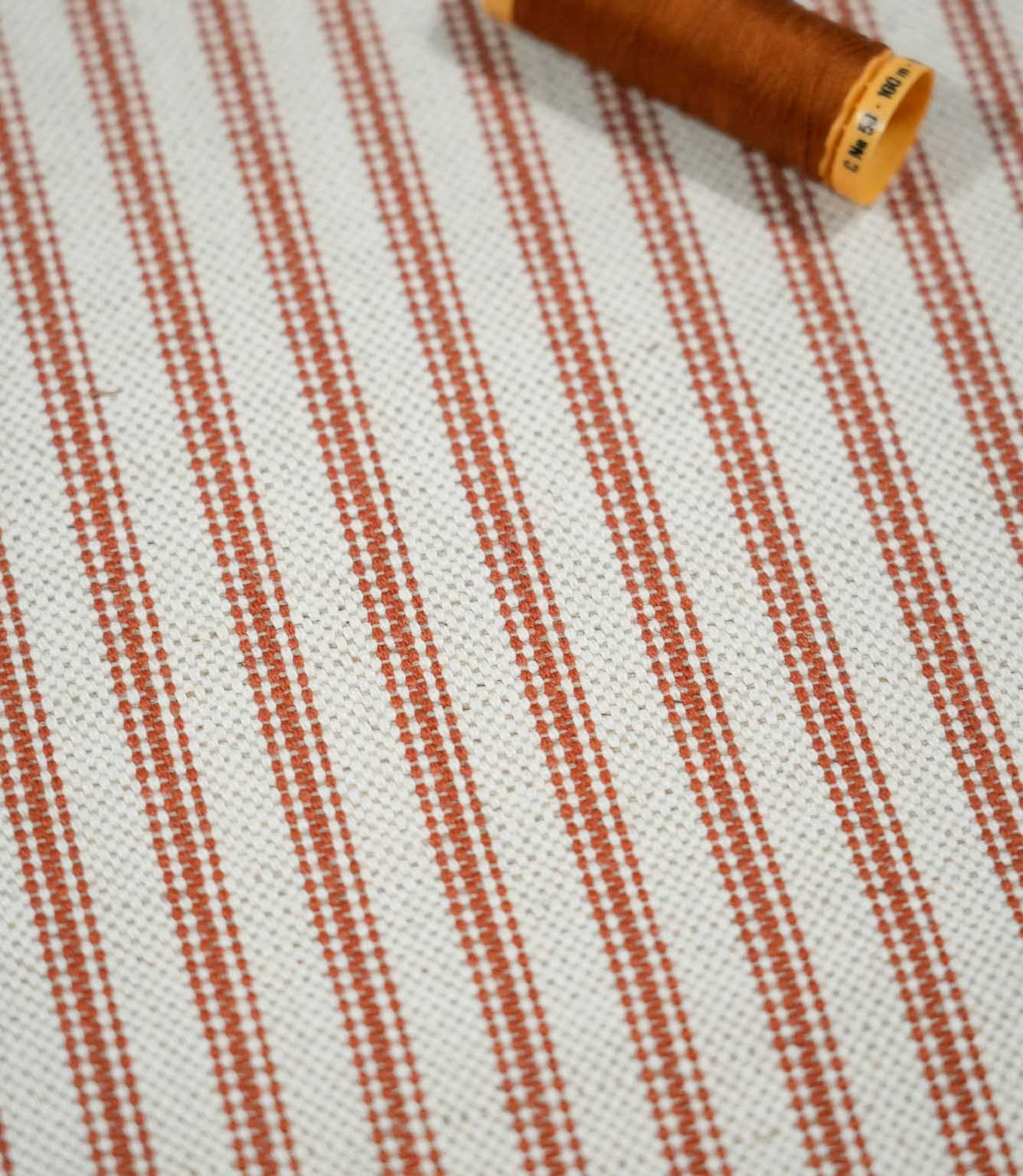 Angled close-up of Linen Ticking Burnt Orange fabric with a textured cream weave, fine burnt orange stripes and coordinating orange thread spool.