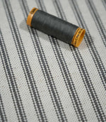 Close-up of grey ticking stripe fabric with a coordinating grey thread spool, highlighting the woven stripe pattern and natural textured finish.