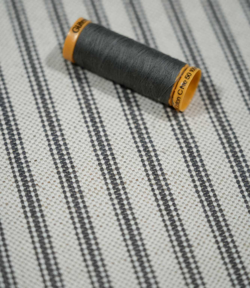 Close-up of grey ticking stripe fabric with a coordinating grey thread spool, highlighting the woven stripe pattern and natural textured finish.