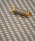 Close-up of natural ticking stripe fabric with a coordinating taupe thread spool, highlighting the ivory stripe pattern and tactile woven texture.