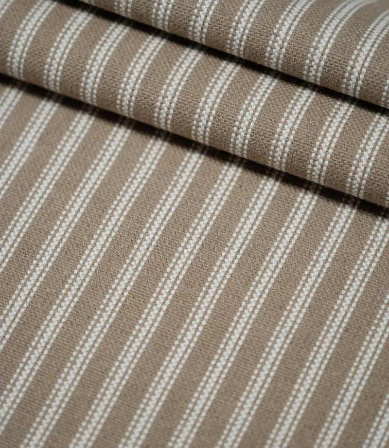 Layered folds of natural ticking stripe fabric with repeating ivory stripes, showing the texture and structure of the warm taupe woven fabric.