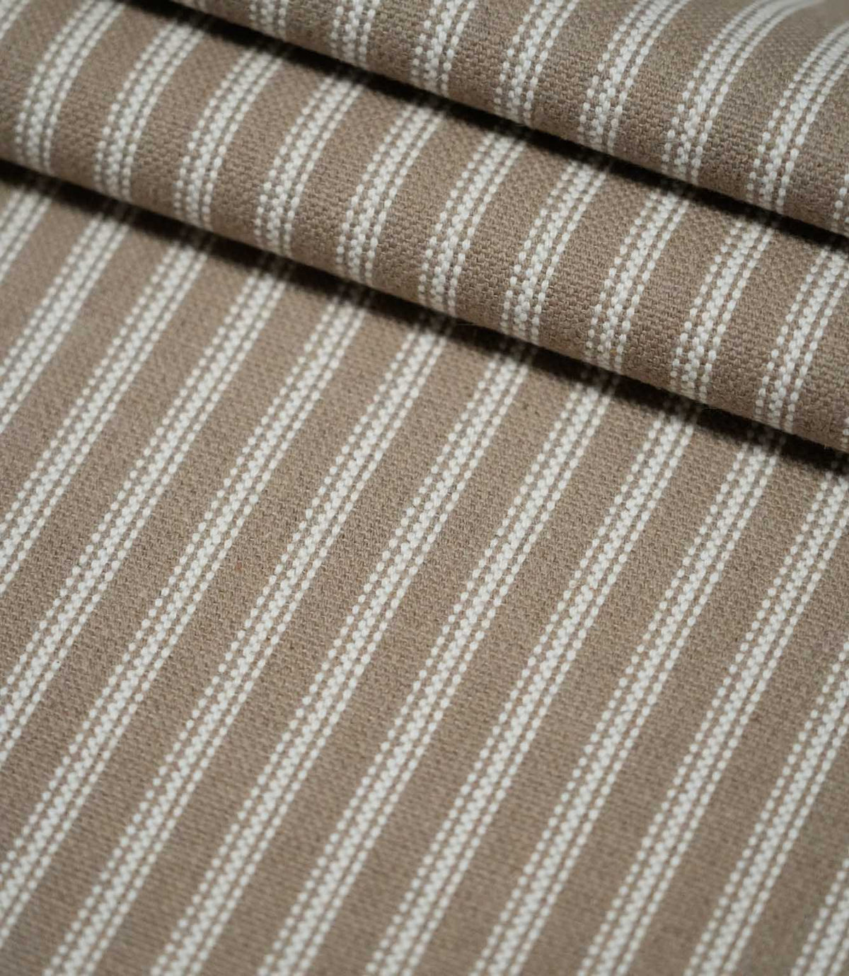 Layered folds of natural ticking stripe fabric with repeating ivory stripes, showing the texture and structure of the warm taupe woven fabric.