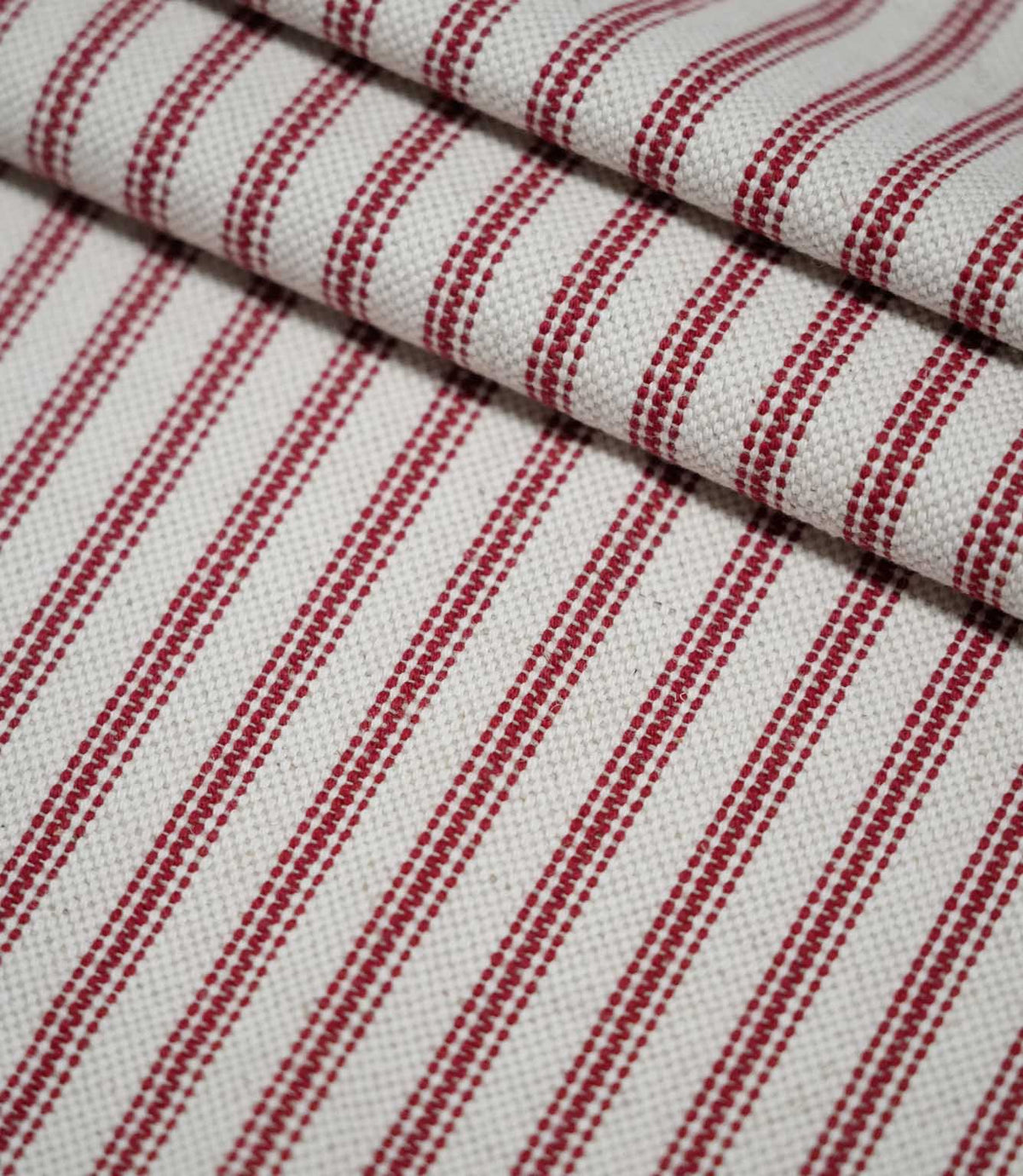 Layered folds of red ticking stripe fabric with evenly spaced red woven stripes on a natural cream background, highlighting the fabric texture and pattern.