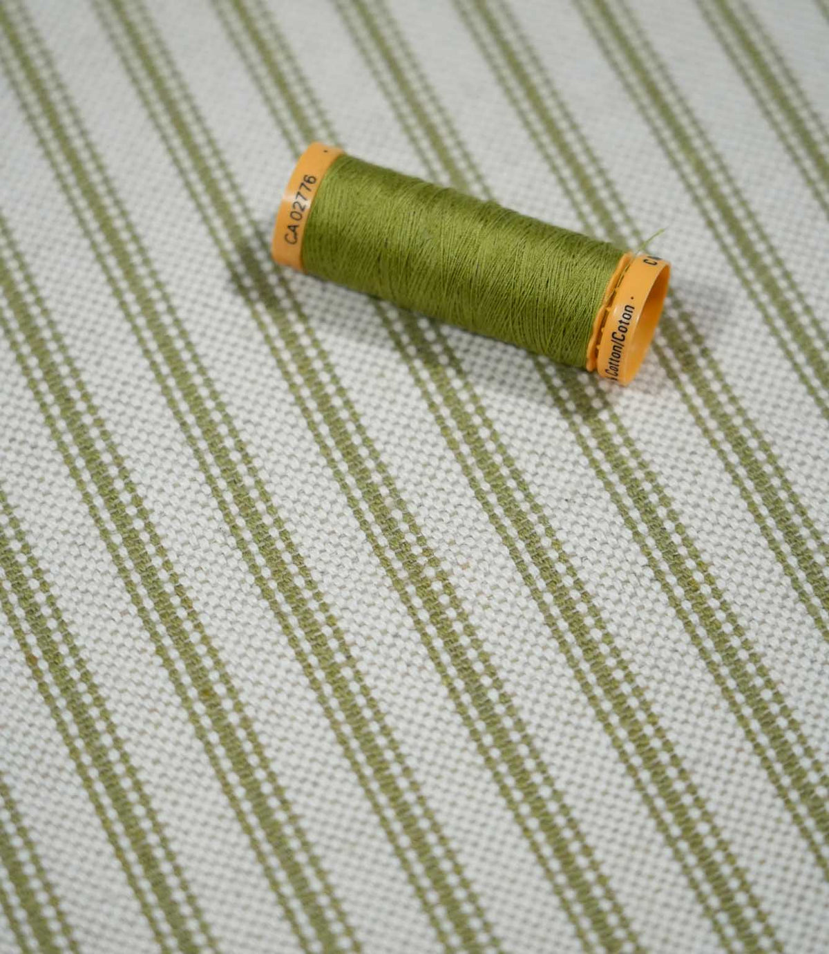 Sage green ticking stripe fabric with a coordinating green thread spool, showing the woven stripe detail and textured natural cream background.