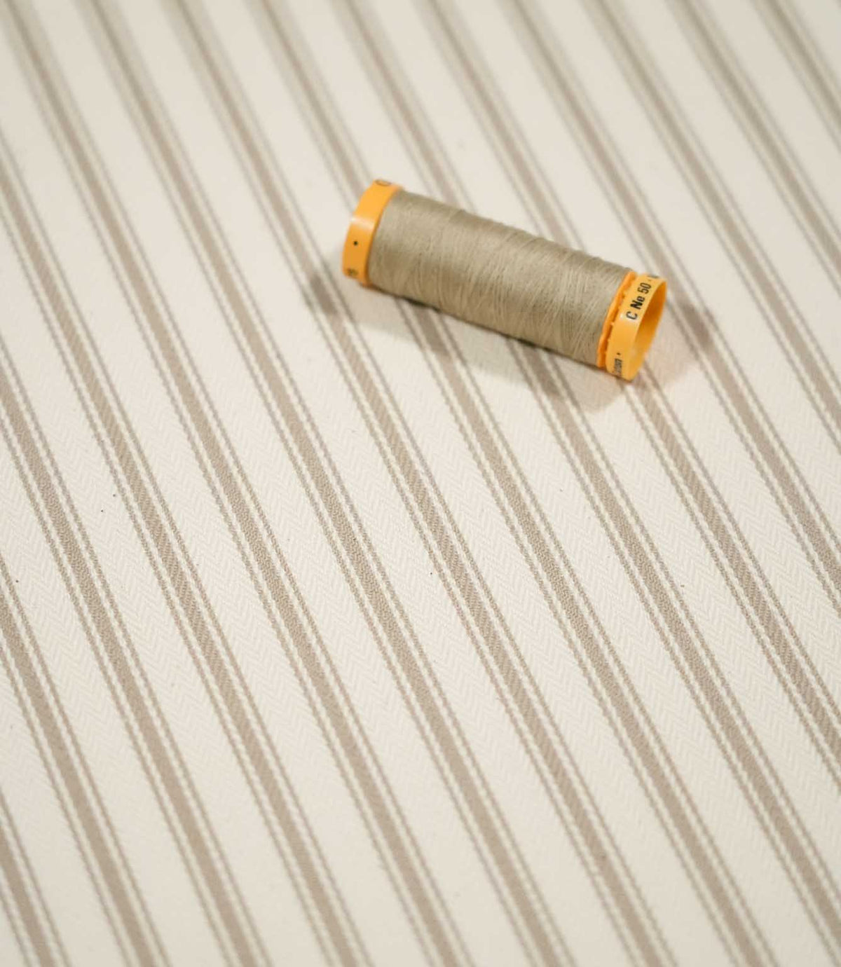 JF Ticking Beige fabric styled with a coordinating beige thread spool, highlighting the classic ticking stripe pattern and subtle woven texture.