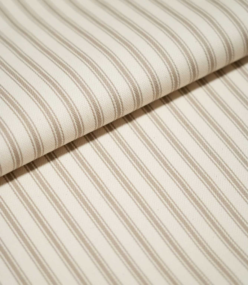 Close-up of folded JF Ticking Beige fabric showing fine beige ticking stripes and a subtle herringbone texture on a warm cream background.