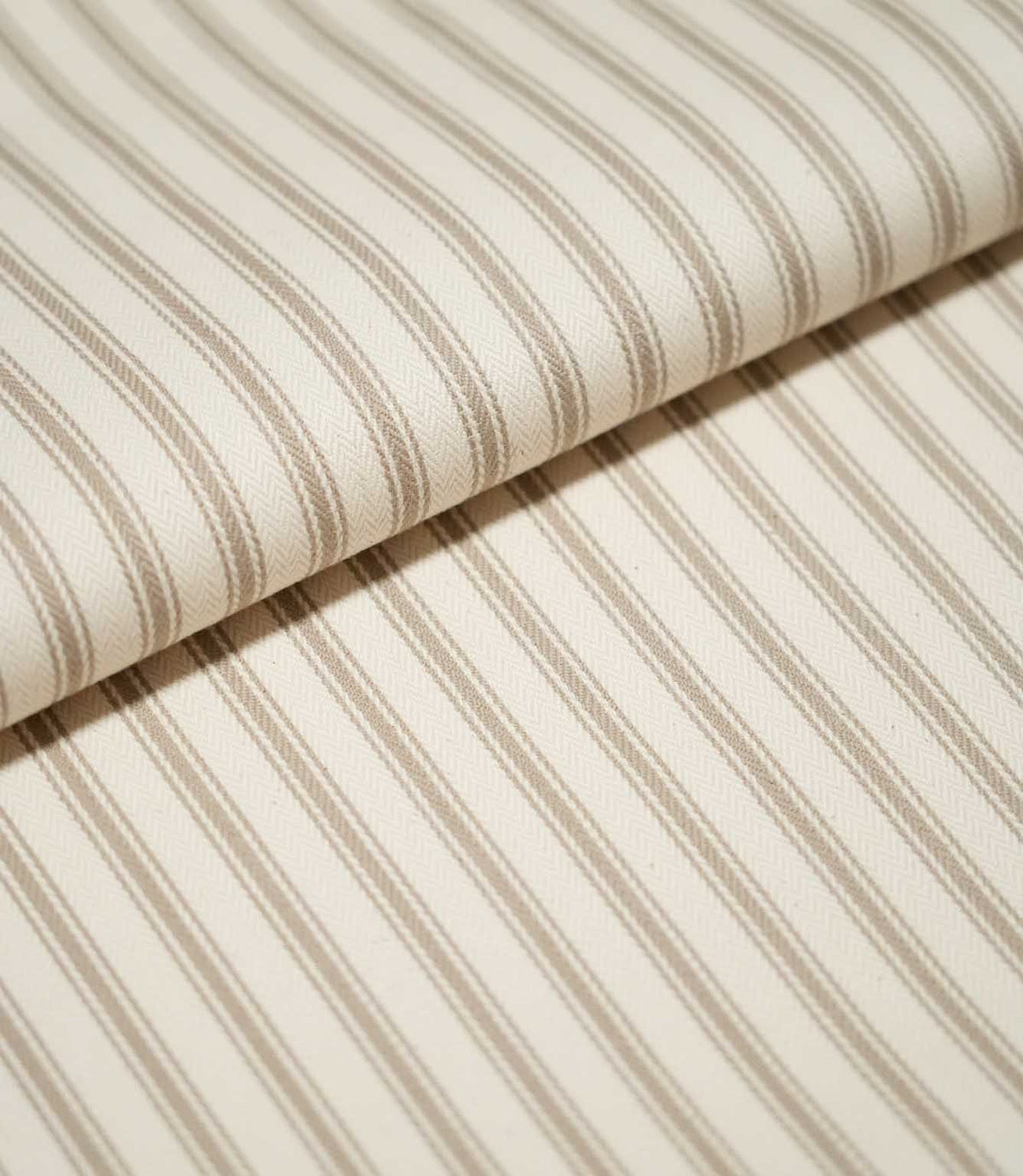 Close-up of folded JF Ticking Beige fabric showing fine beige ticking stripes and a subtle herringbone texture on a warm cream background.