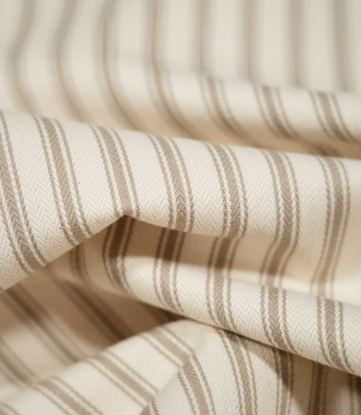 Close-up of layered JF Ticking Beige fabric showing the natural drape, cream herringbone weave and repeating fine beige ticking stripes.