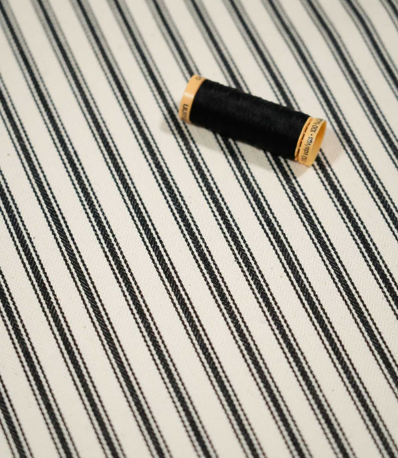 JF Ticking Black fabric styled with a coordinating black thread spool, highlighting the classic stripe pattern and subtle woven herringbone texture.
