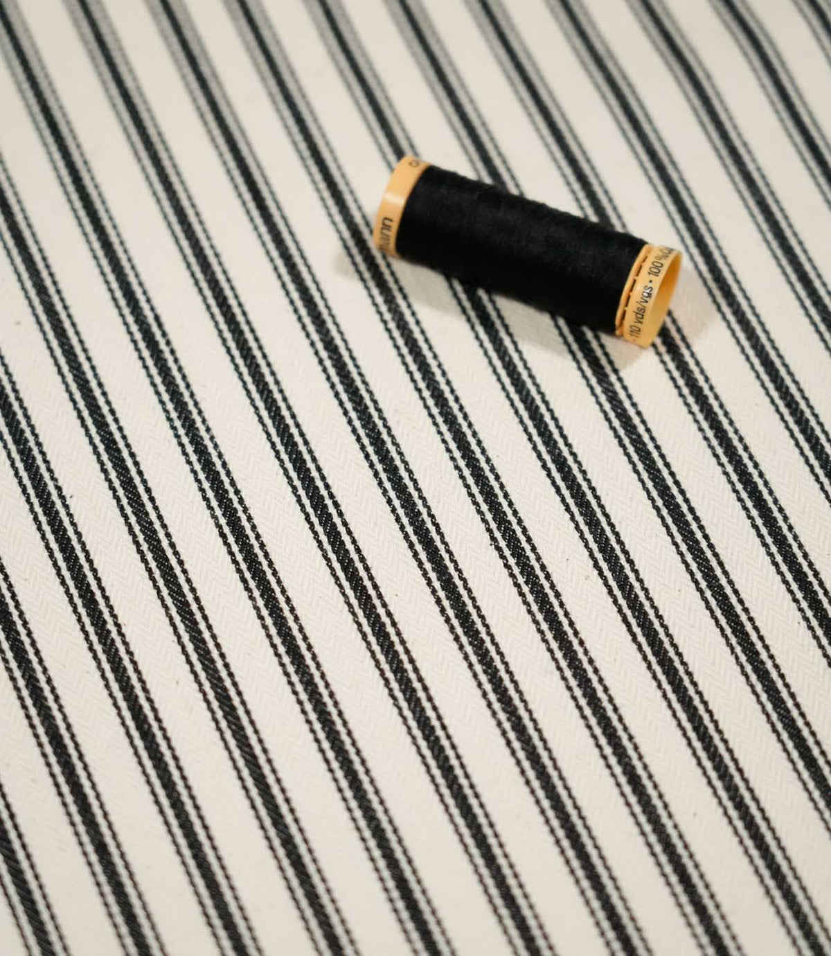 JF Ticking Black fabric styled with a coordinating black thread spool, highlighting the classic stripe pattern and subtle woven herringbone texture.