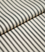 Close-up of folded JF Ticking Black fabric showing crisp black ticking stripes and a subtle herringbone weave on a warm cream background.