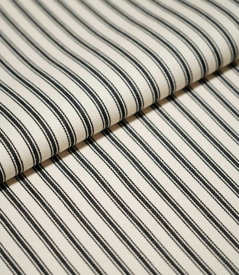 Close-up of folded JF Ticking Black fabric showing crisp black ticking stripes and a subtle herringbone weave on a warm cream background.
