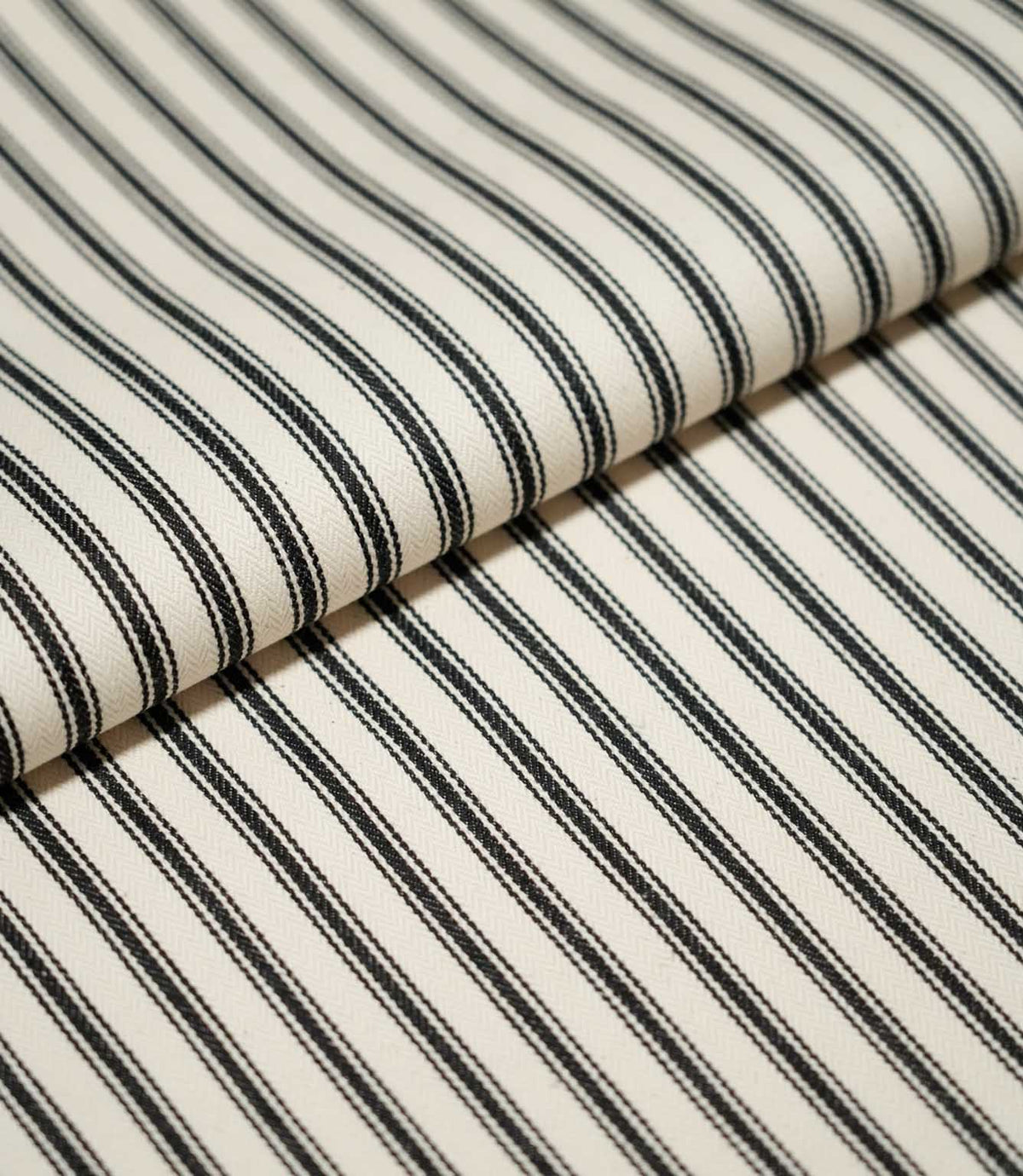 Close-up of folded JF Ticking Black fabric showing crisp black ticking stripes and a subtle herringbone weave on a warm cream background.