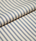 Close-up of folded JF Ticking Blue fabric showing the repeating blue stripe pattern and subtle textured herringbone weave on a cream background.