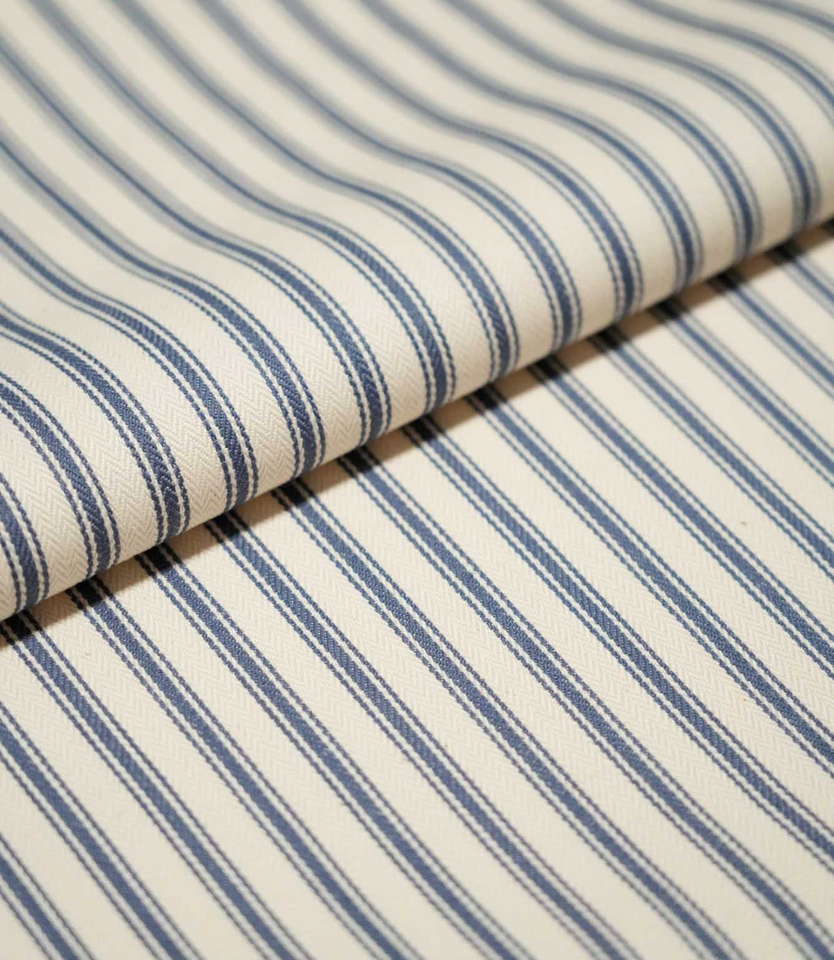 Close-up of folded JF Ticking Blue fabric showing the repeating blue stripe pattern and subtle textured herringbone weave on a cream background.