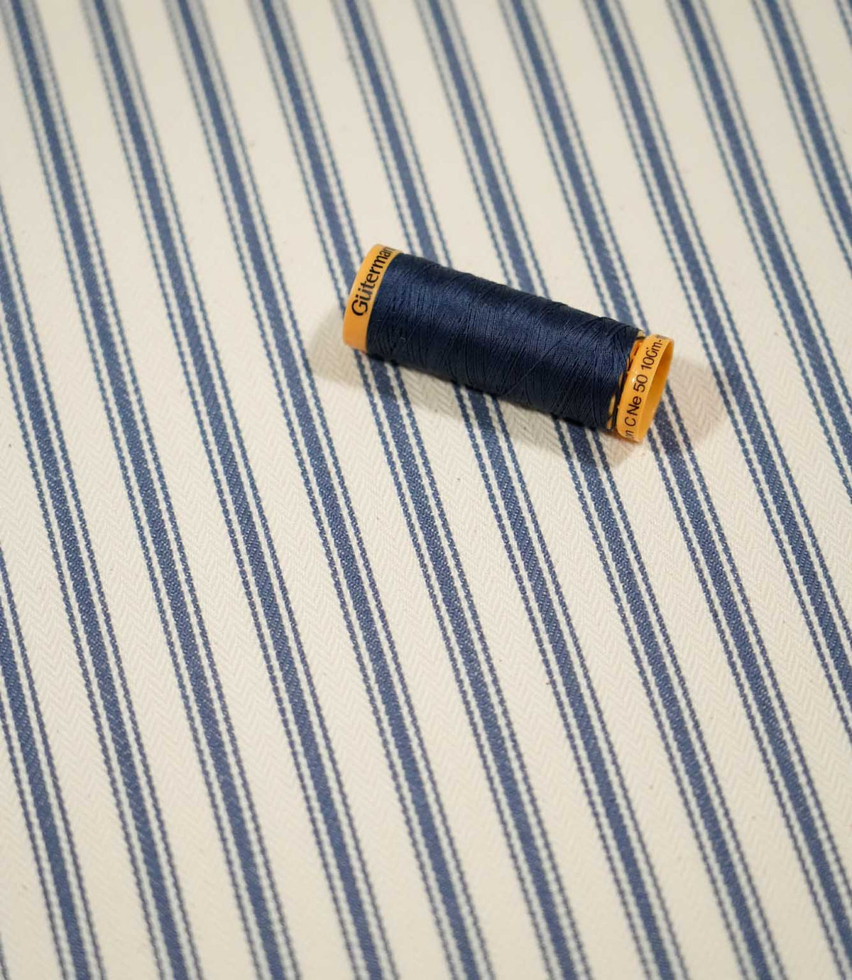 JF Ticking Blue fabric styled with a coordinating navy thread spool, highlighting the classic blue ticking stripes and cream woven ground.